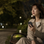 夜の公園のベンチでスマートフォンを握り既婚者との恋に悩む日本人女性